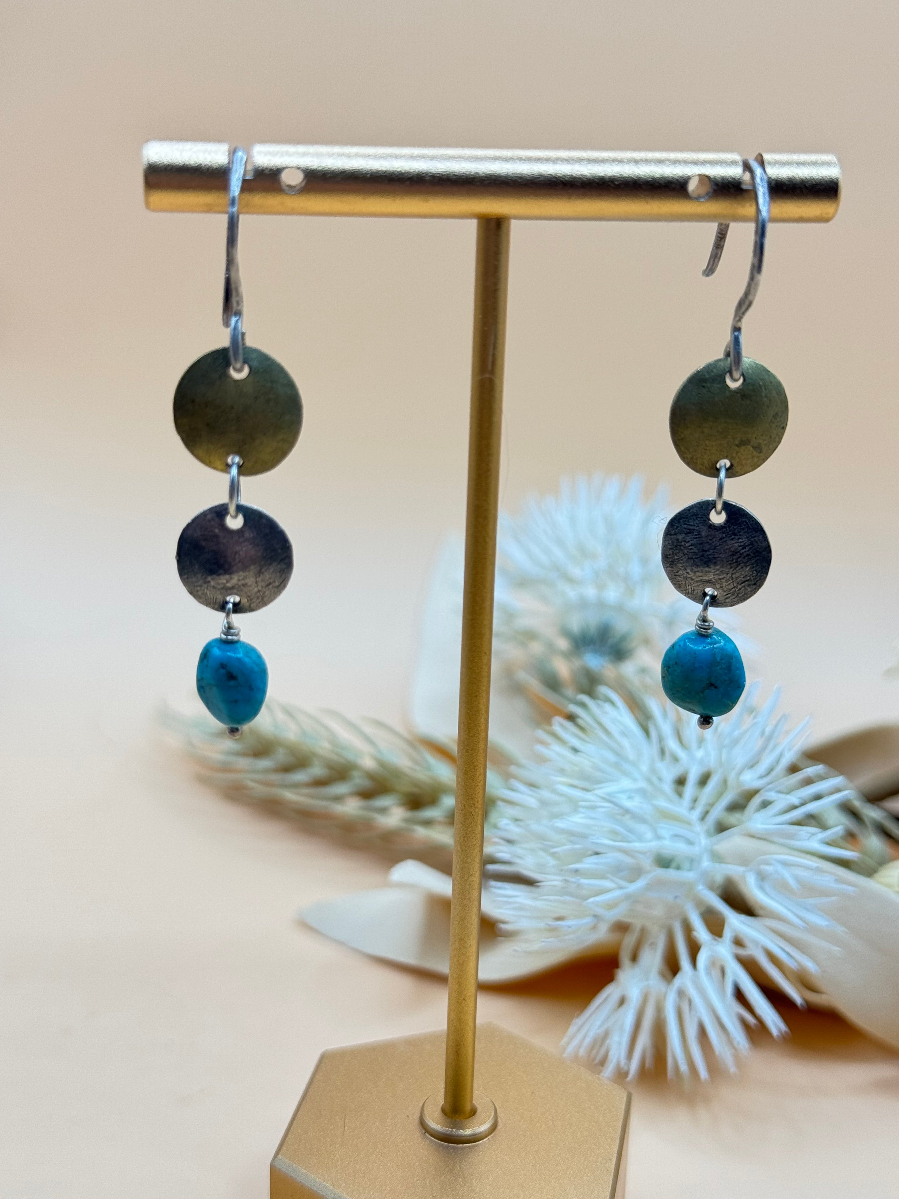 Artisan mixed metal dangle earrings featuring brass and silver tone disc accents with vibrant turquoise nugget beads. Their handcrafted boho style makes them a unique accessory for everyday wear or Southwestern-inspired looks.